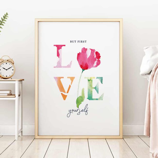 Ein Bilderrahmen steht an eine Zimmerwand geleht auf einem Dielenboden. Das Motiv ist ein farbenfrohe Collage aus dem Wort LOVE und einer Blüte. Der Text darauf lautet „But first LOVE yourself“